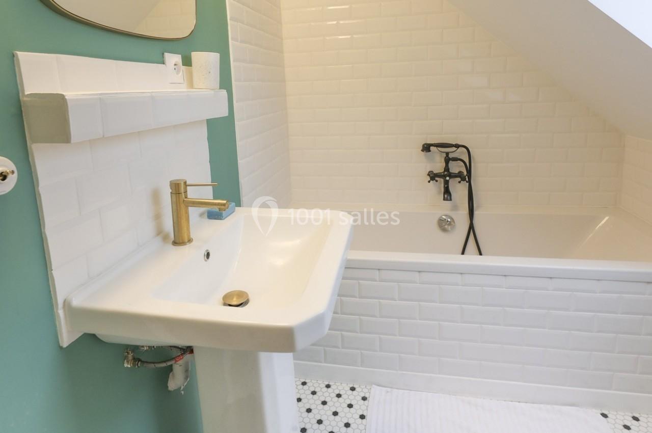 Salle de bain avec un lavabo blanc, un robinet doré, une baignoire et des murs en carrelage blanc.