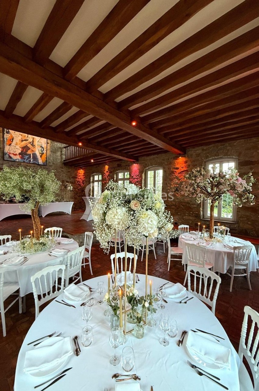 Salle de réception avec tables rondes décorées de fleurs blanches et chandeliers, sous un plafond en poutres apparentes.