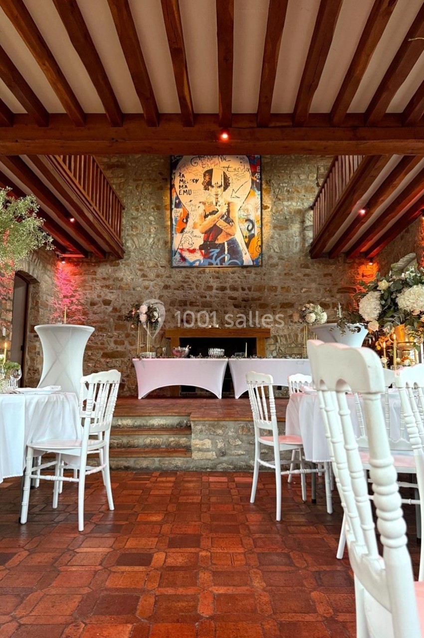 Salle de réception avec murs en pierre, plafond en bois, tables décorées et tableau coloré en arrière-plan.