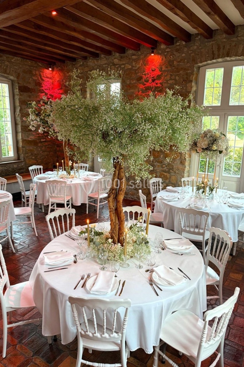 Salle de réception avec tables rondes dressées, nappes blanches et centre de table végétal sous un plafond en bois.