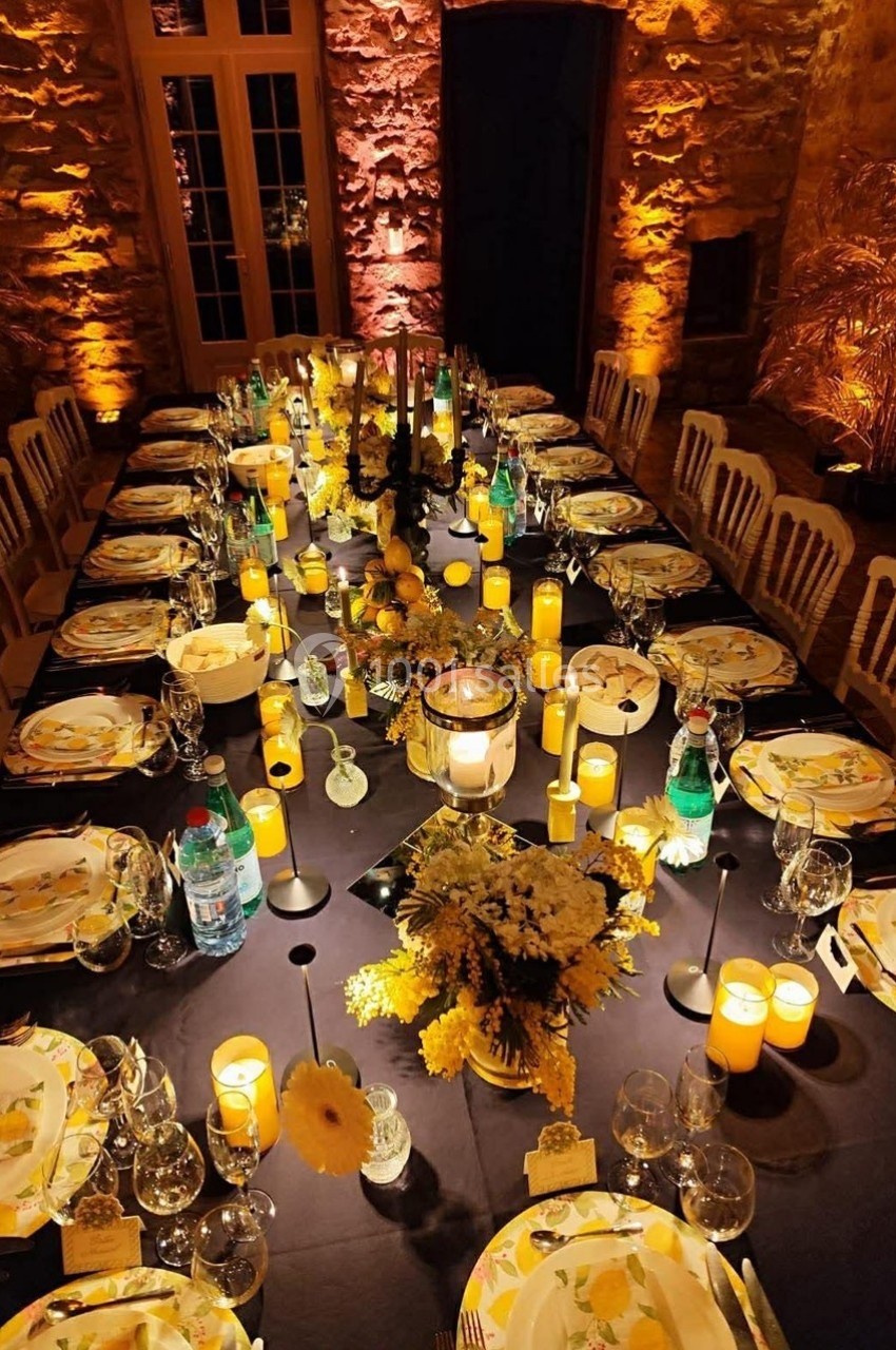 Table élégamment dressée avec des assiettes décorées, bougies allumées et fleurs jaunes dans une salle en pierre éclairée.