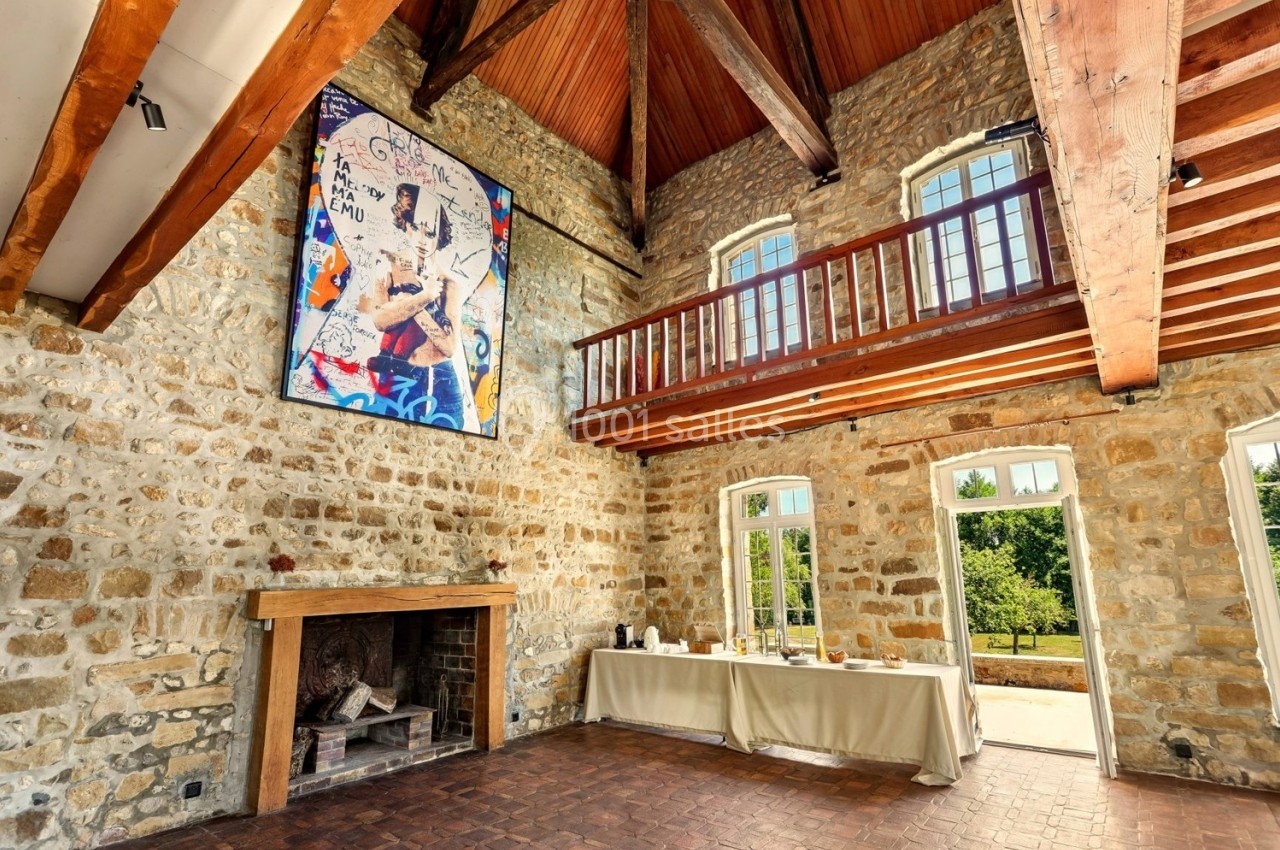 Salle en pierre avec cheminée, mezzanine en bois, tableau coloré au mur et table dressée près de grandes fenêtres.