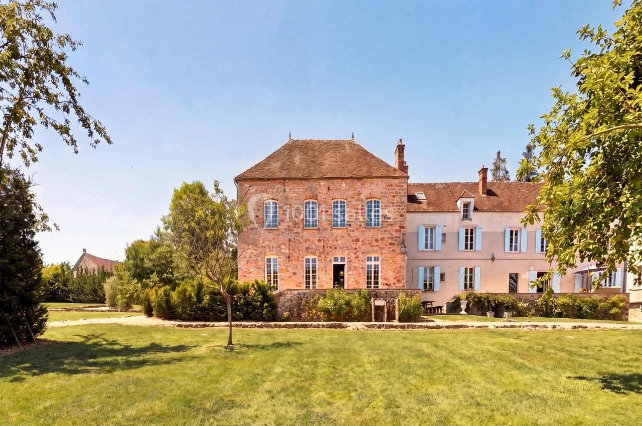 Grande maison en pierre et crépi rose avec volets bleus, entourée d'un jardin verdoyant sous un ciel dégagé.