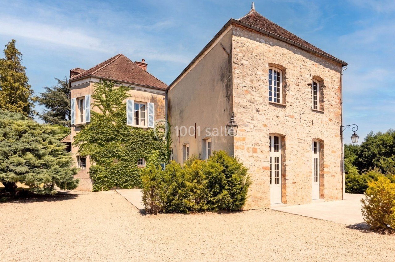 Bâtiment en pierre avec toit en pente, volets blancs, lierre grimpant et allée gravillonnée sous un ciel bleu.