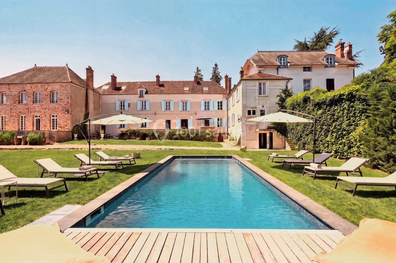 Piscine extérieure entourée de chaises longues et parasols, avec des bâtiments anciens en arrière-plan.