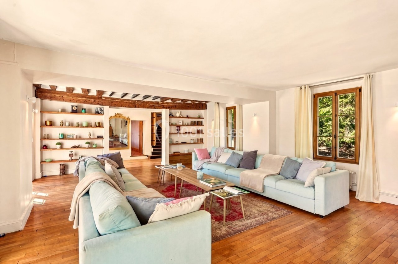 Salon lumineux avec parquet en bois, canapés pastel, tapis rouge, étagères décorées et grandes fenêtres donnant sur l…