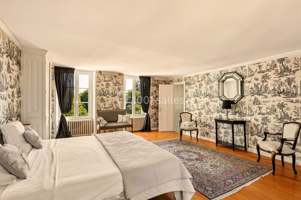 Chambre lumineuse avec lit double, papier peint à motifs, tapis coloré, fauteuils et vue sur un jardin par les fenêtres.