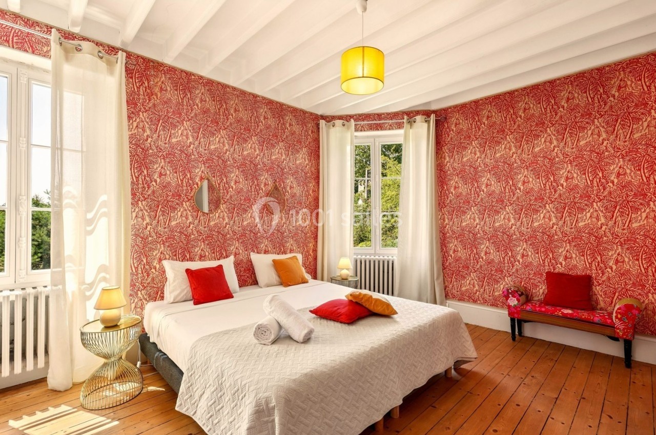 Chambre lumineuse avec murs rouges à motifs, lit double, coussins colorés, parquet en bois et grandes fenêtres.