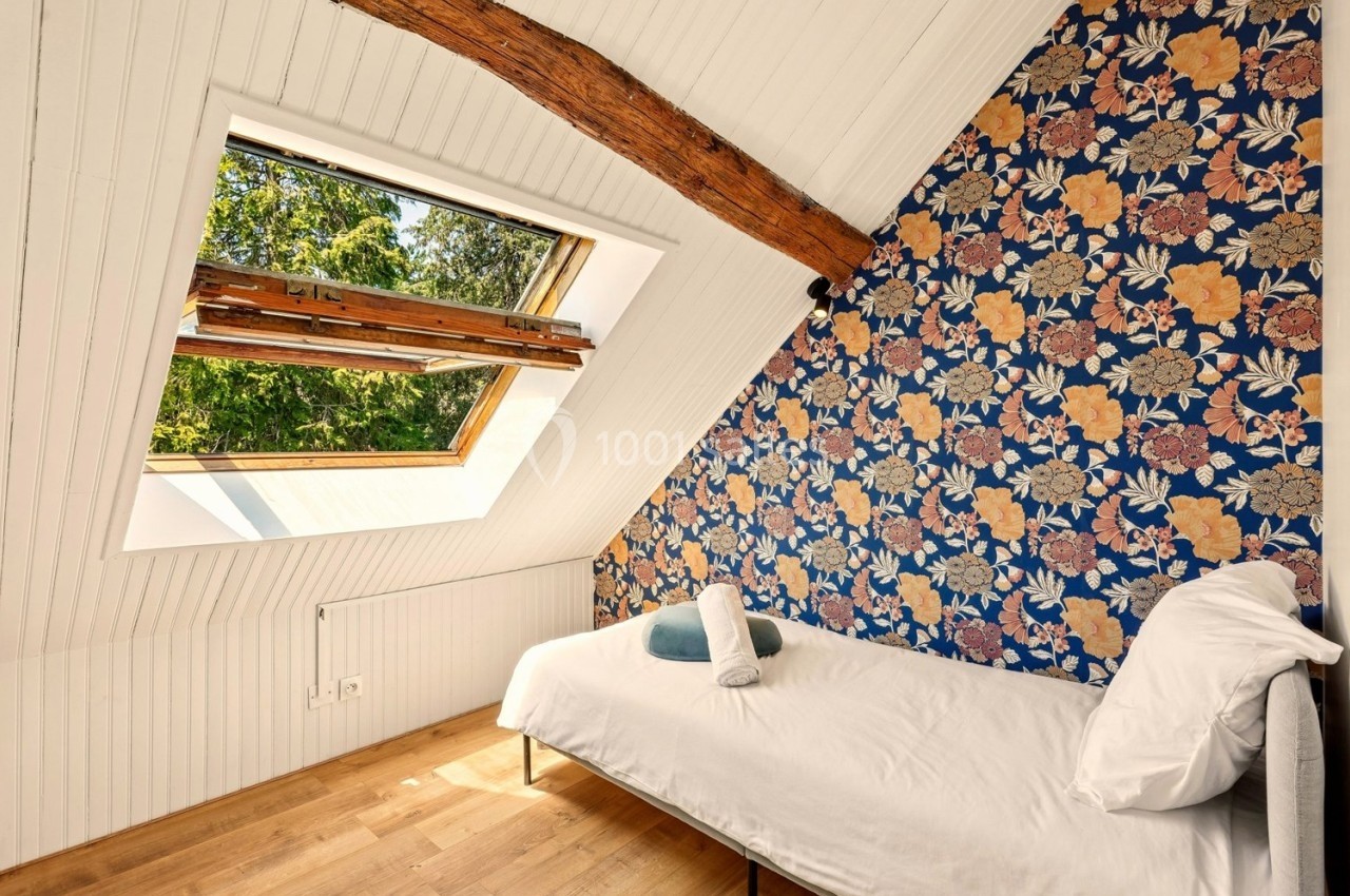 Chambre mansardée avec lit simple, mur fleuri, poutre apparente et fenêtre de toit donnant sur des arbres.