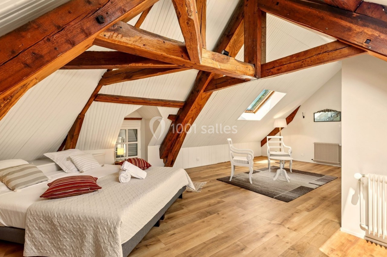 Chambre mansardée avec poutres apparentes, lit double, fauteuils blancs, parquet et lumière naturelle d'une fenêtre de toit.