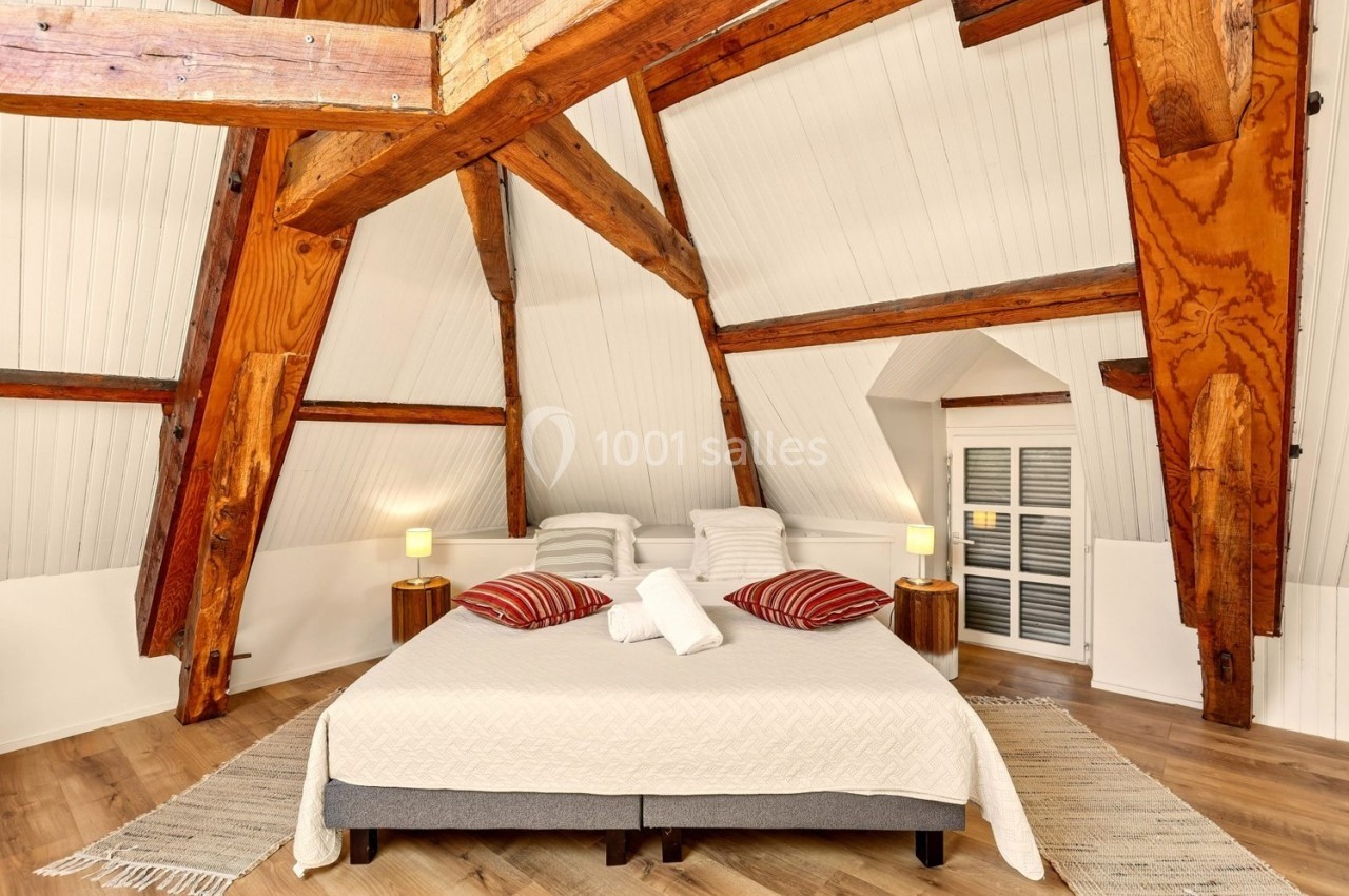 Chambre mansardée avec poutres apparentes, lit double, coussins rouges, lampes de chevet et parquet clair.