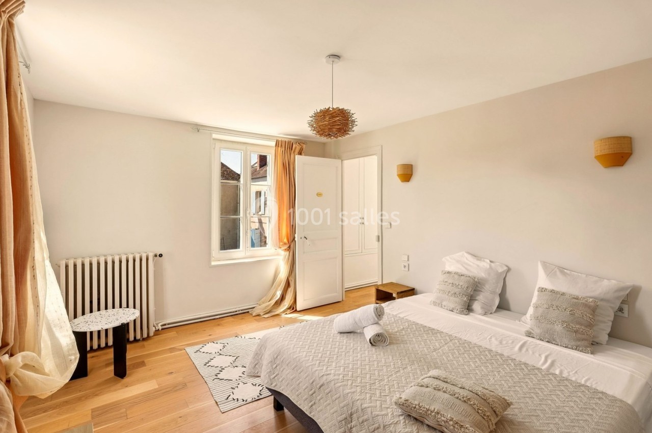 Chambre lumineuse avec lit double, parquet, rideaux beige clair, et vue sur des bâtiments par une fenêtre ouverte.