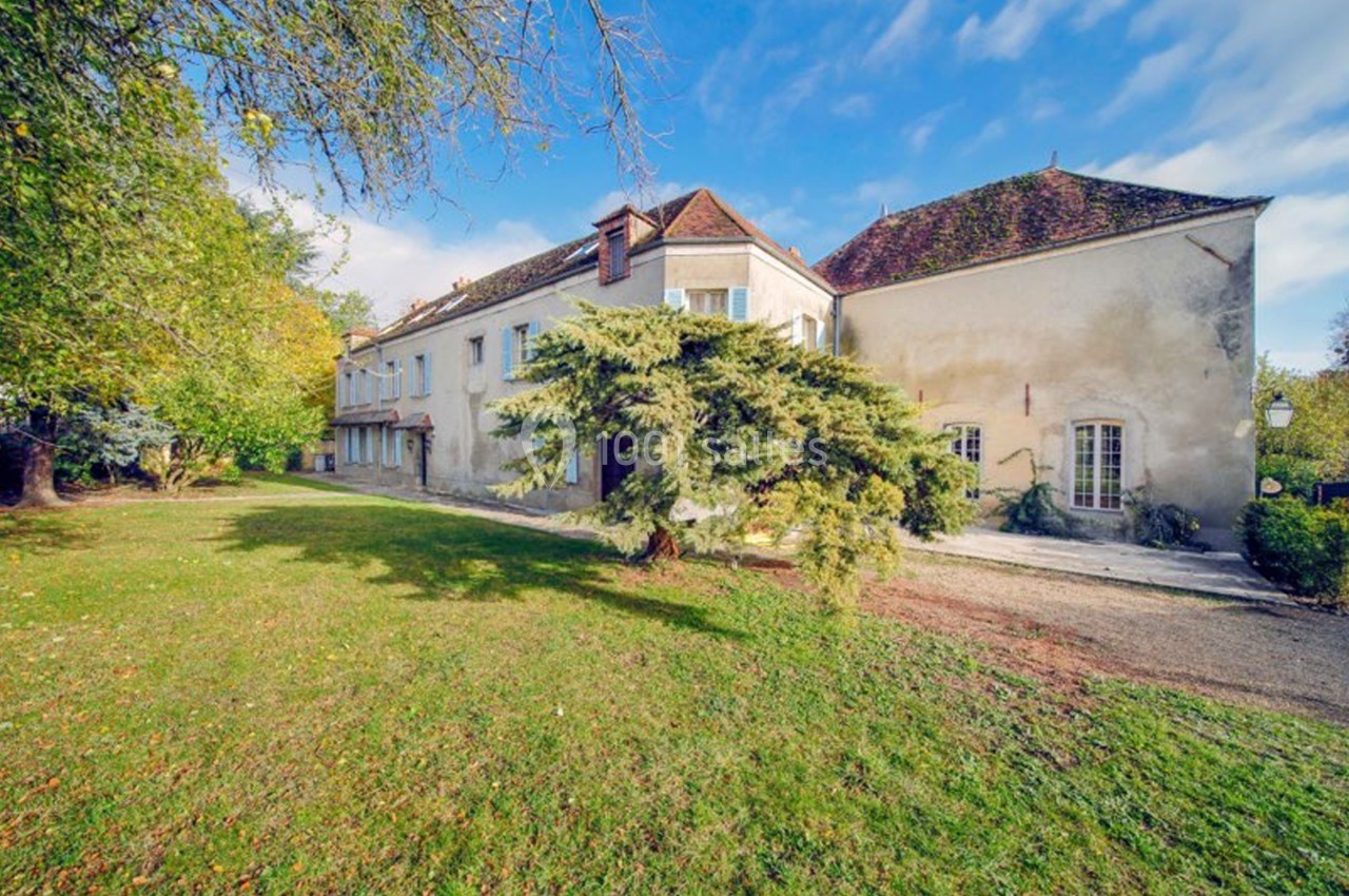 Grande maison ancienne avec toiture en tuiles, entourée d'un jardin arboré sous un ciel partiellement nuageux.