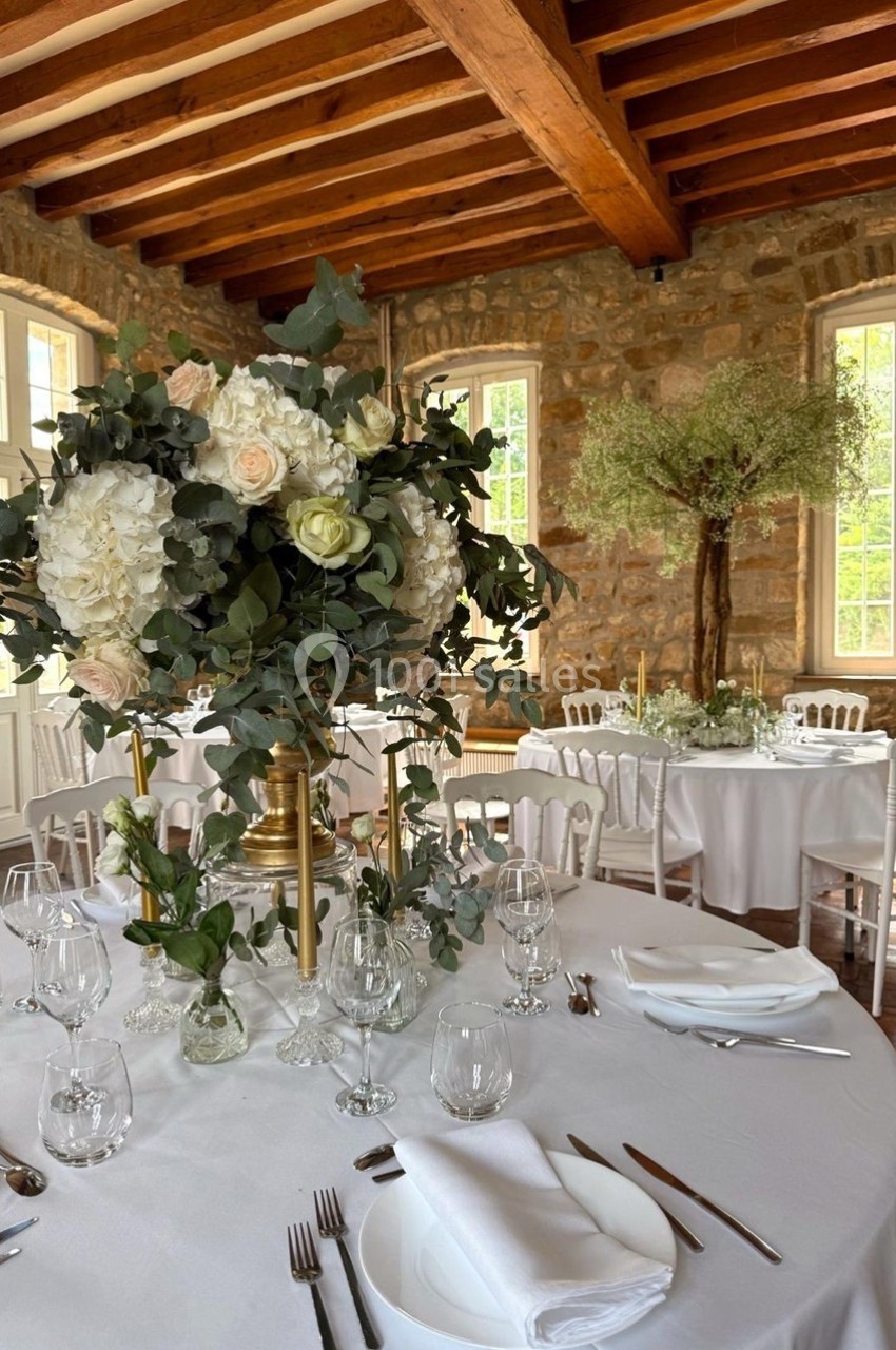 Salle de réception lumineuse avec tables rondes dressées, décor floral élégant et poutres apparentes au plafond.
