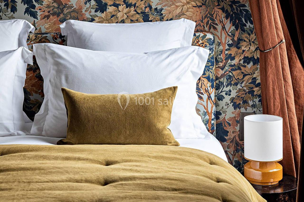 Lit avec coussins blancs et oreillers moutarde, devant une tête de lit à motif floral, avec une lampe sur une table de…