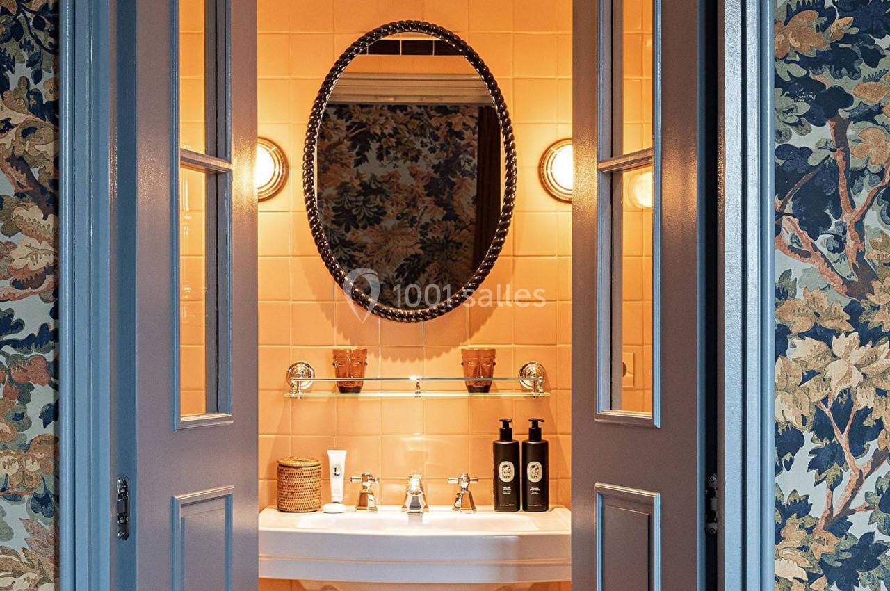 Lavabo avec miroir ovale, éclairage mural et décor floral sur les murs et carrelage beige en arrière-plan.