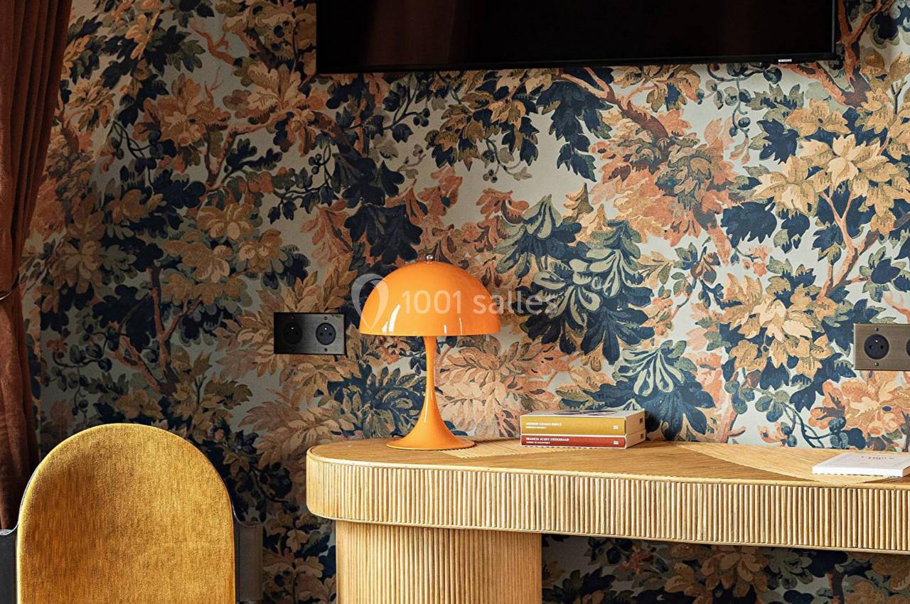 Lampe orange sur un bureau en bois, avec un fond de papier peint floral et des rideaux marron.