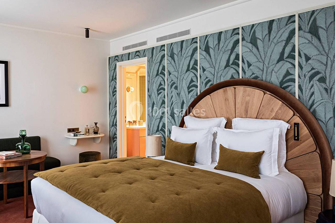 Chambre d'hôtel avec lit double, tête de lit en bois, décor mural à motifs végétaux et coin salon avec table ronde.