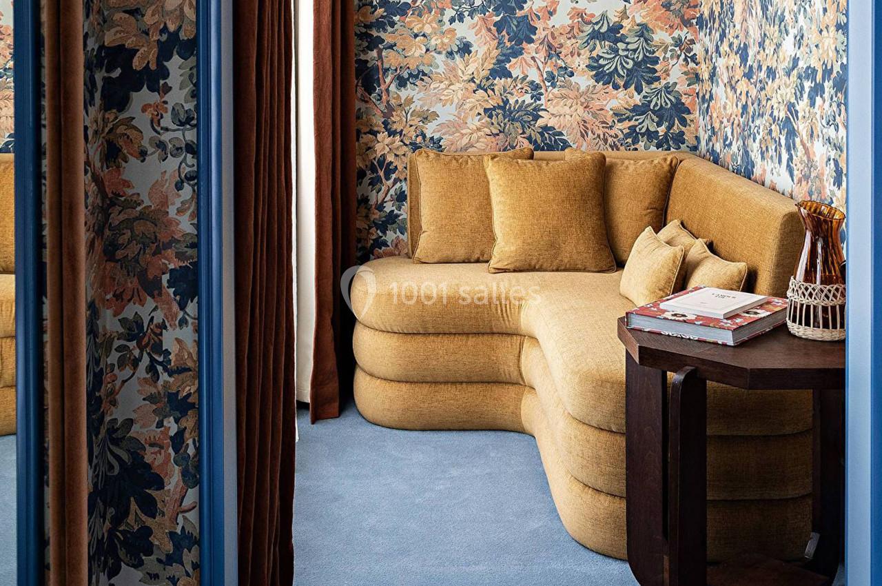 Canapé d'angle jaune moutarde dans une pièce aux murs tapissés de motifs floraux, avec une table en bois à côté.