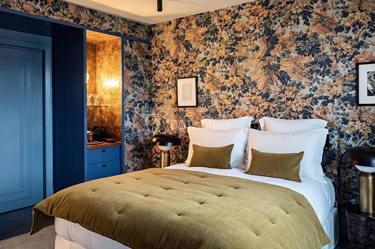 Chambre élégante avec lit double, couvre-lit vert olive, murs à motifs floraux et éclairage tamisé.