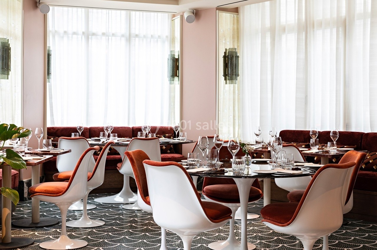 Salle de restaurant lumineuse avec tables dressées, chaises blanches et banquettes rouges, décorée de plantes et de rideaux.