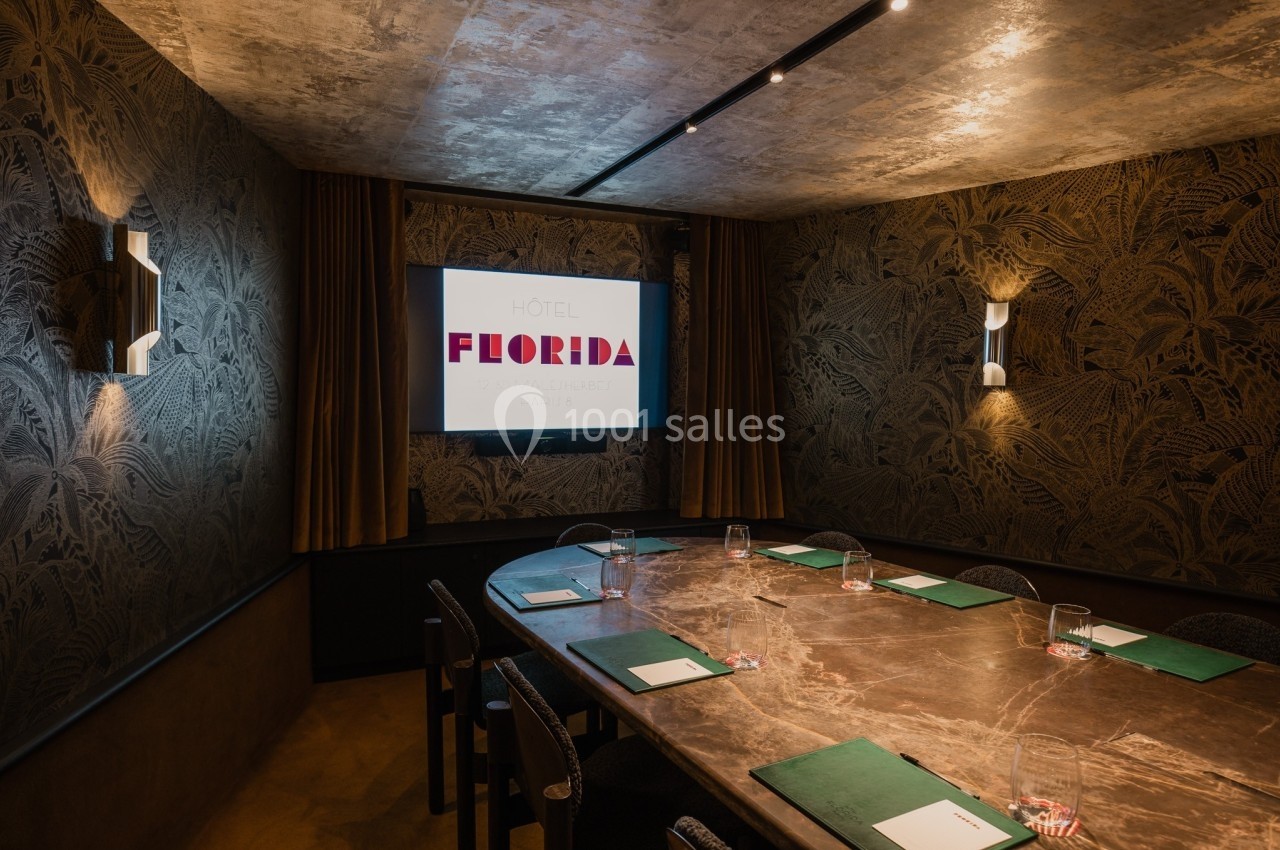 Salle de réunion avec une grande table ovale en marbre, écran de présentation allumé et murs décorés de motifs végétaux.