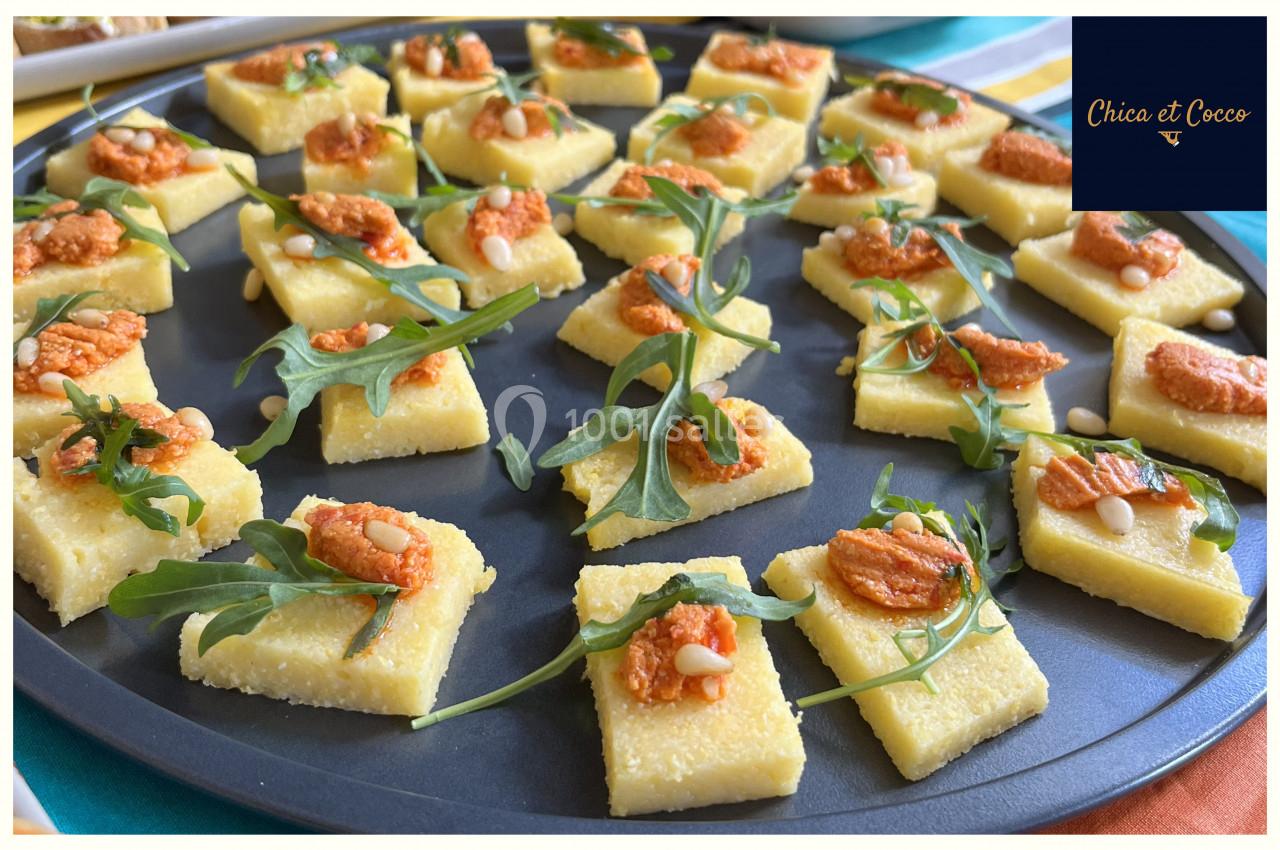 Carrés de polenta garnis de crème rouge, roquette et pignons, disposés sur un plateau rond.