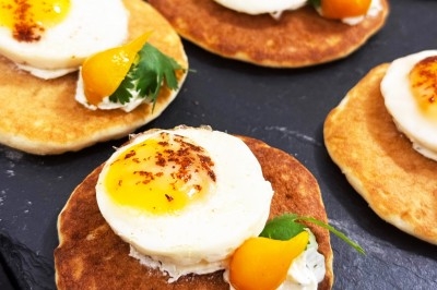 Mini-blinis garnis d'œufs au plat, crème et herbes fraîches, présentés sur une surface sombre.