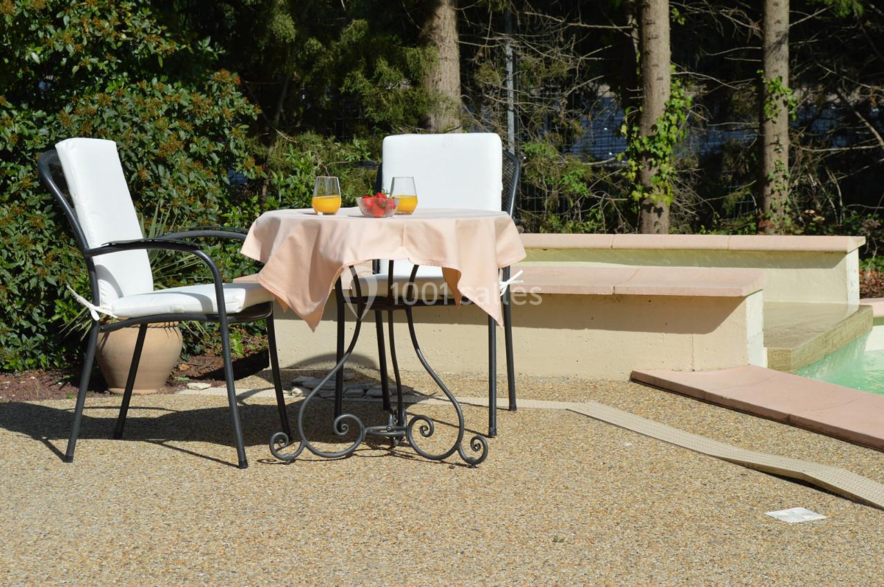 Table ronde avec nappe beige, deux chaises blanches, verres de jus d'orange et fraises, disposée près d'une piscine.