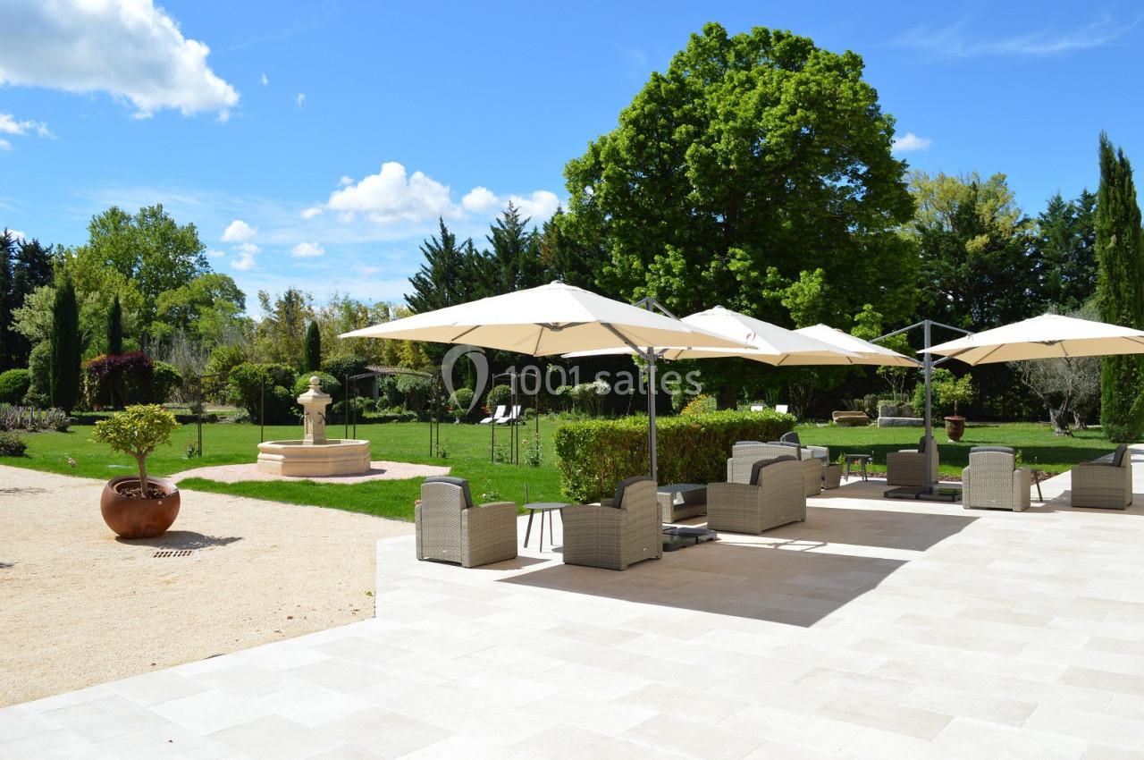 Terrasse avec fauteuils et parasols donnant sur un jardin verdoyant, parsemé d'arbres et d'une fontaine centrale.