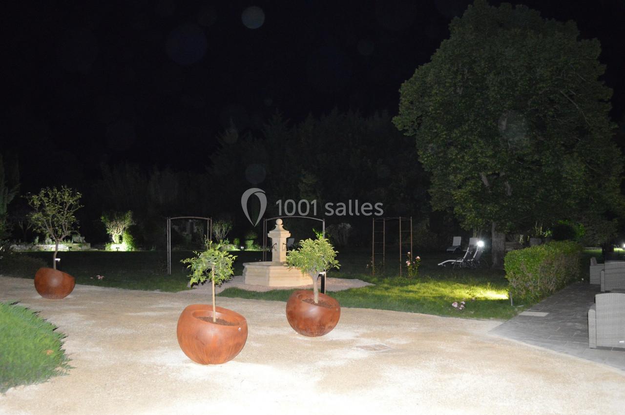 Allée de jardin éclairée la nuit, avec des pots en métal contenant de petits arbres et une fontaine en arrière-plan.