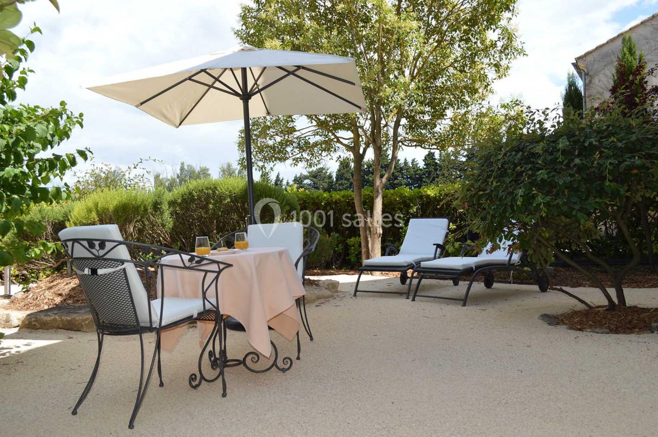 Table de jardin avec nappe beige, chaises en métal, parasol blanc et transats sur une terrasse entourée de verdure.