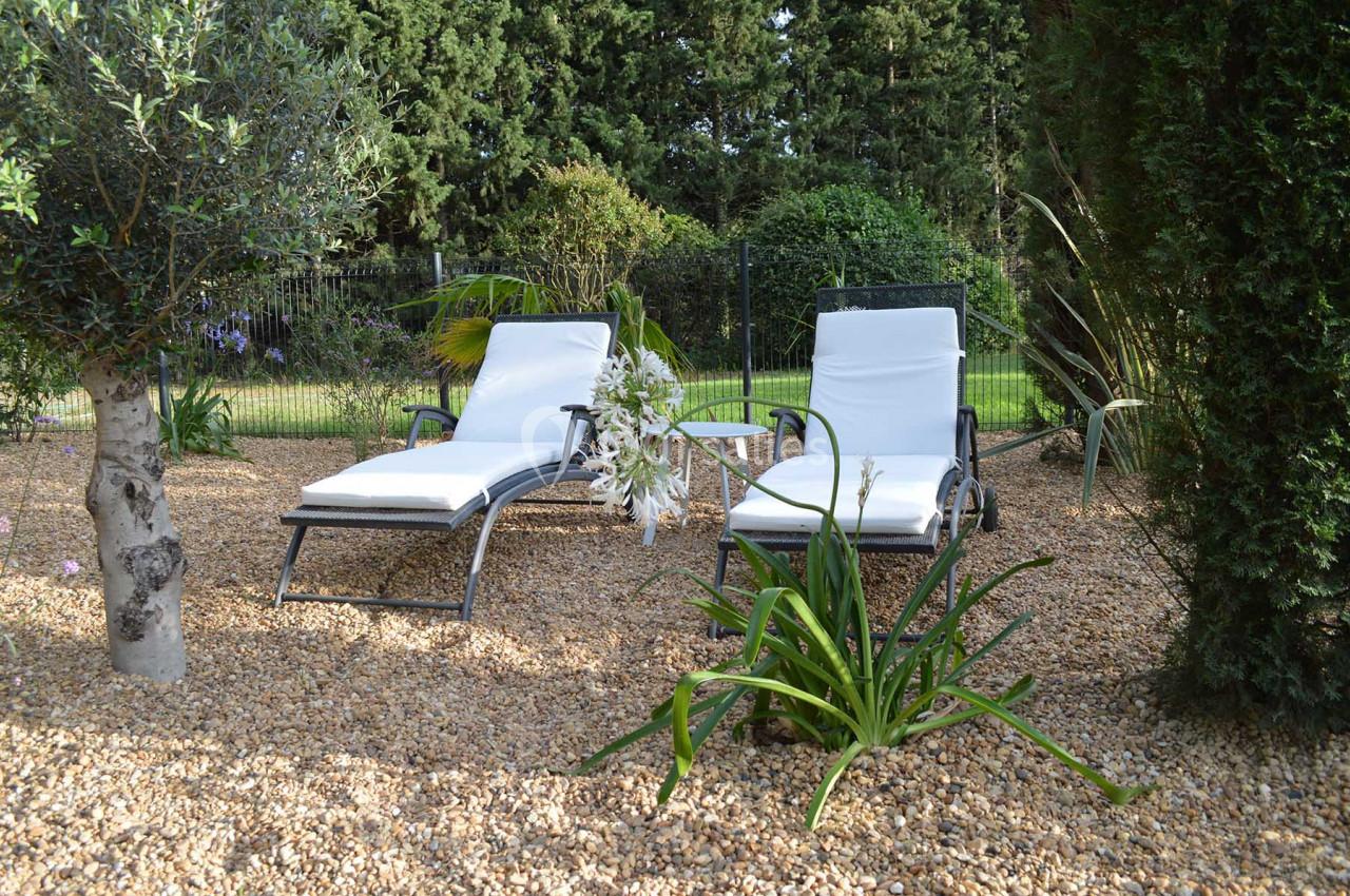 Deux chaises longues blanches sur un sol en gravier, entourées de plantes et d'arbres dans un jardin.