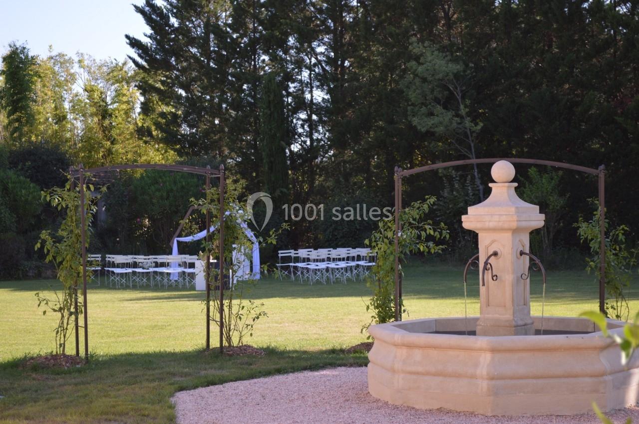 Fontaine en pierre au premier plan dans un jardin verdoyant, avec des chaises blanches disposées sous une arche au fond.