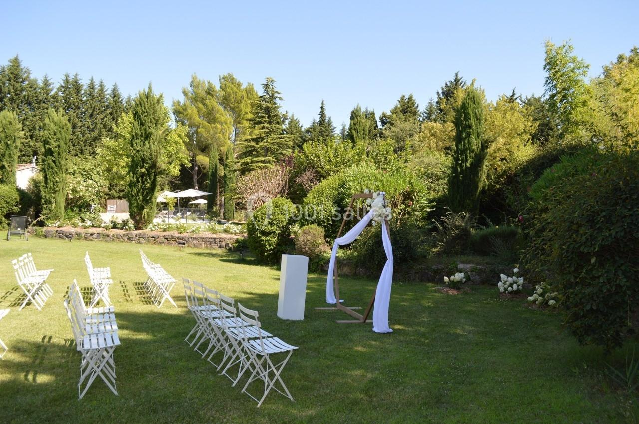 Chaises blanches disposées en extérieur devant une arche décorée, entourées de verdure et d'arbres.