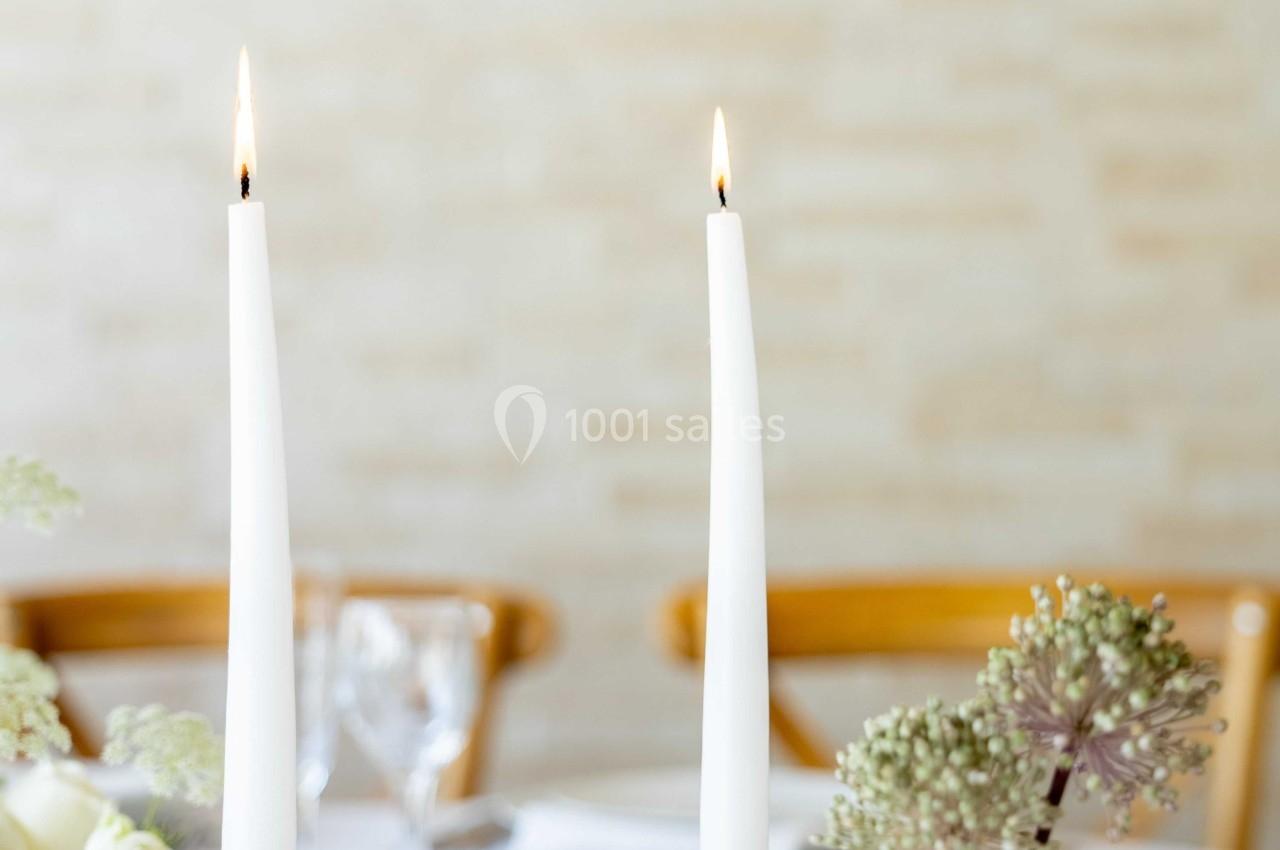 Deux bougies blanches allumées sur des chandeliers, avec un vase de fleurs et une table dressée en arrière-plan.
