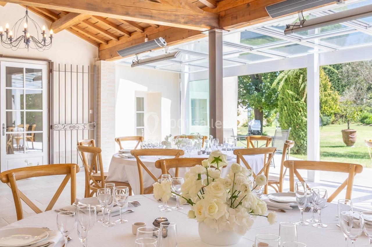Salle lumineuse avec tables dressées, nappes blanches, vaisselle élégante, bouquet de fleurs et plafond en bois.