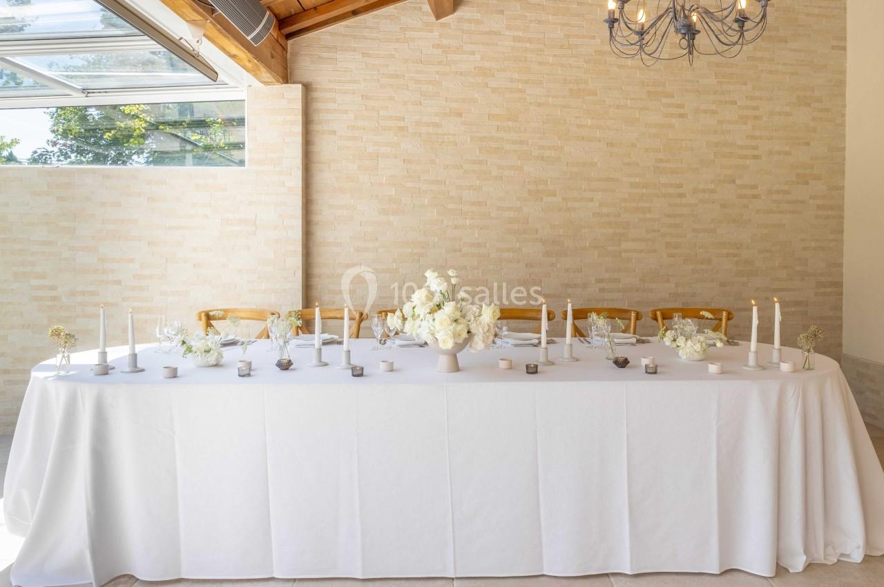 Table décorée avec une nappe blanche, des bougies, des fleurs blanches et des chaises en bois dans une salle lumineuse.