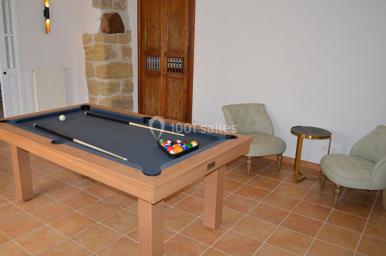 Table de billard en bois avec tapis noir, billes et queue, dans une pièce avec sol en carrelage et fauteuils.