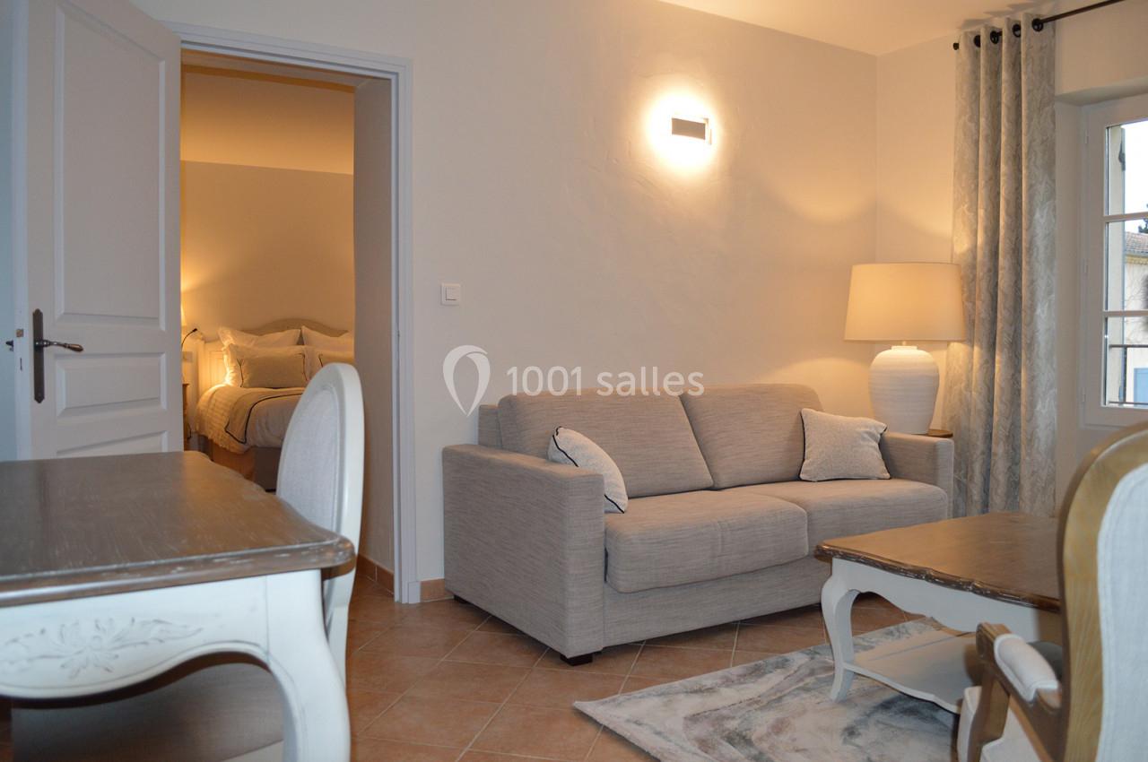 Salon lumineux avec canapé beige, table basse en bois, lampadaire, et vue sur une chambre avec lit double.