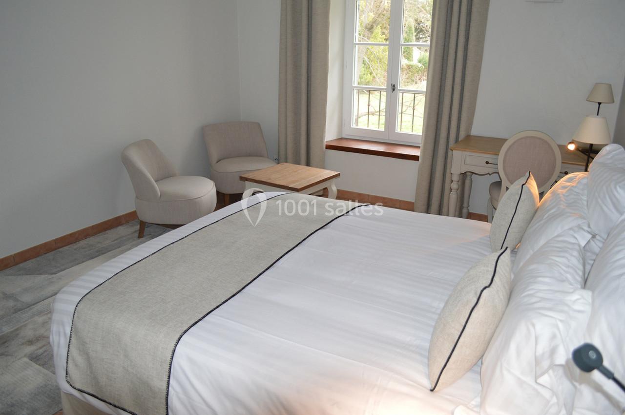 Chambre lumineuse avec lit double, bureau, fauteuils et table basse, près d'une fenêtre donnant sur un jardin.