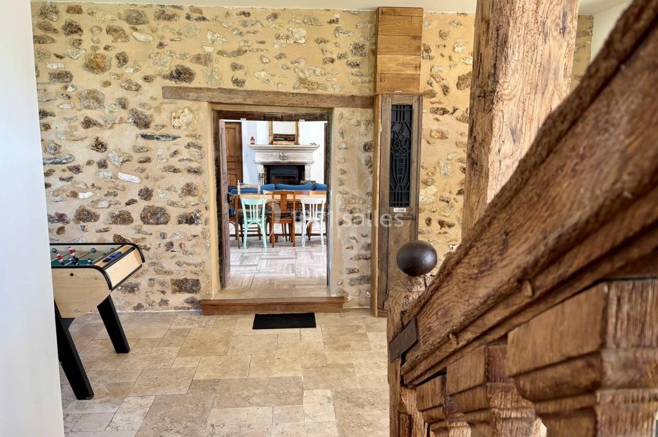 Vue intérieure d'une maison en pierre avec un baby-foot, un escalier en bois et une ouverture vers une salle à manger.