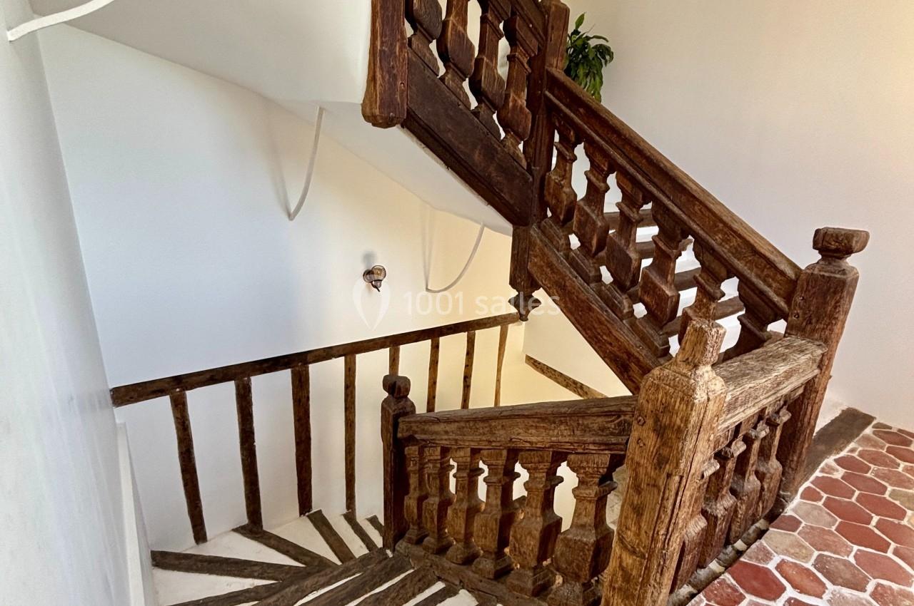 Escalier en bois ancien avec rambardes sculptées, murs blancs et sol en carreaux de terre cuite.