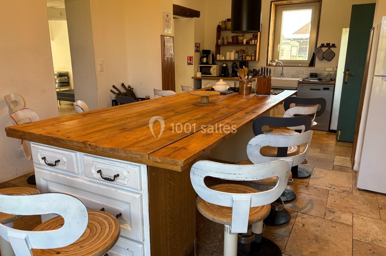 Cuisine lumineuse avec une grande table en bois, des chaises blanches, des étagères et des appareils électroménagers.