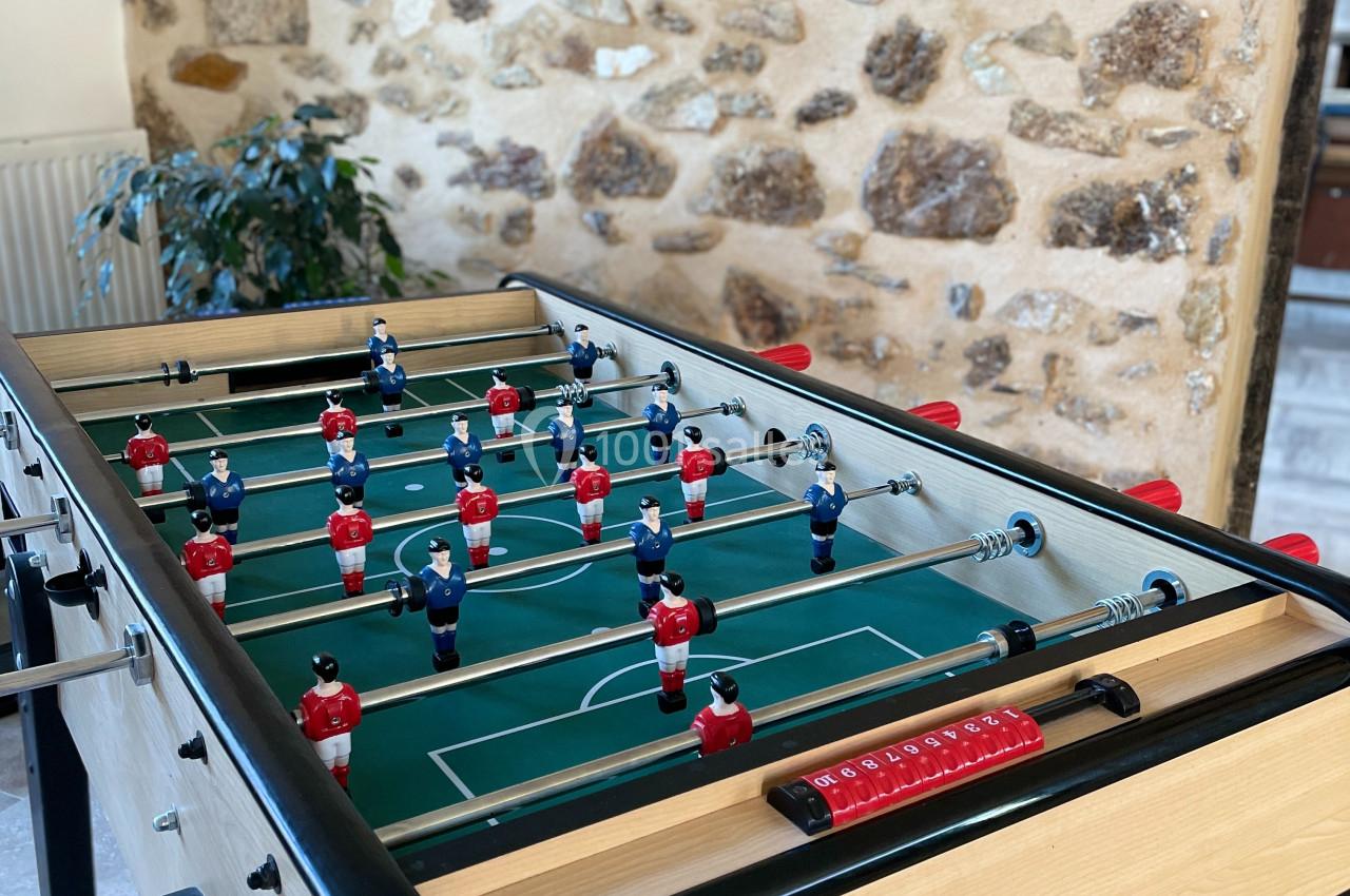 Table de baby-foot en bois clair avec figurines rouges et bleues, placée dans une pièce aux murs en pierre apparente.