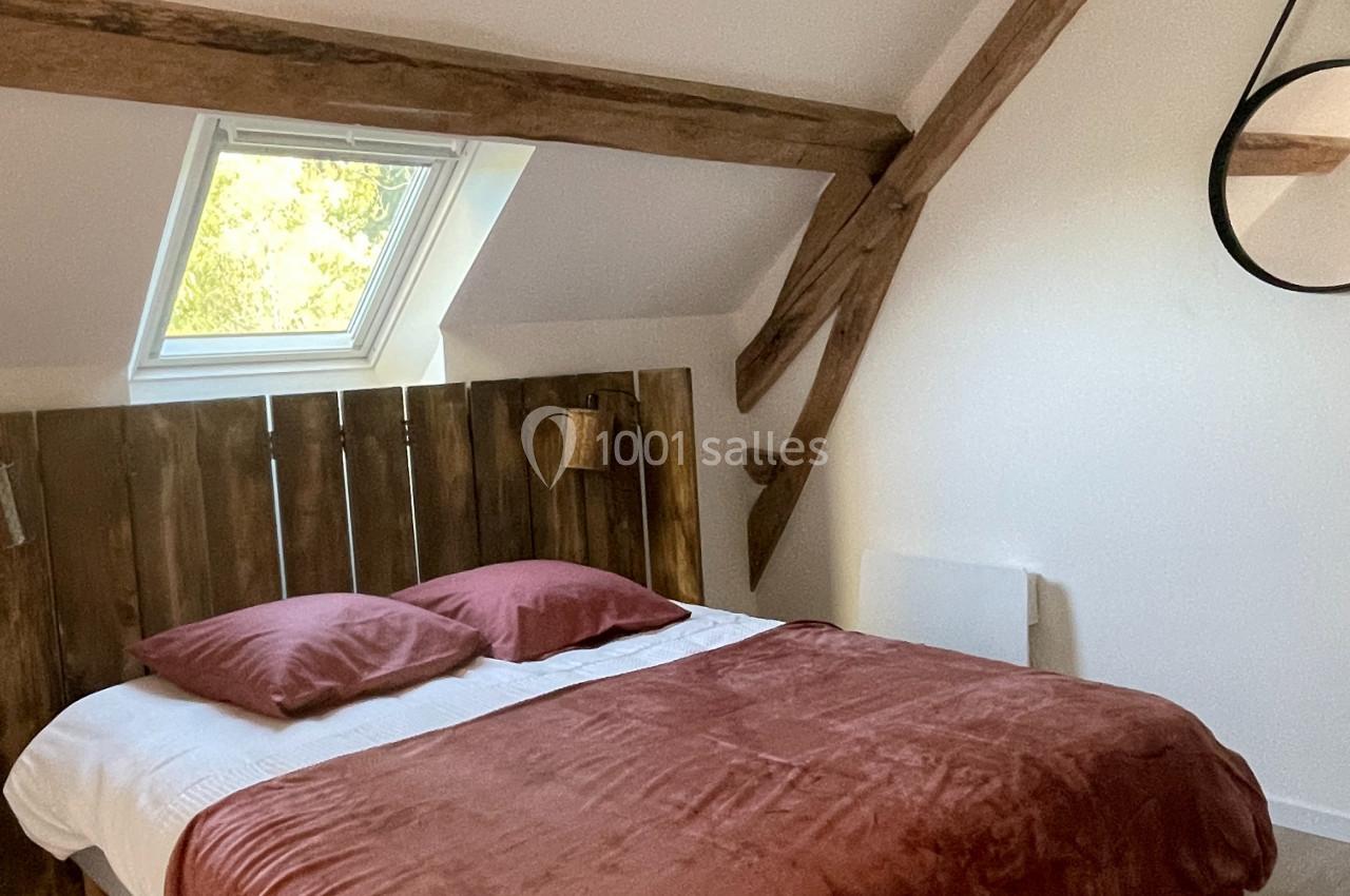 Chambre lumineuse avec lit double, tête de lit en bois, poutres apparentes et fenêtre de toit.