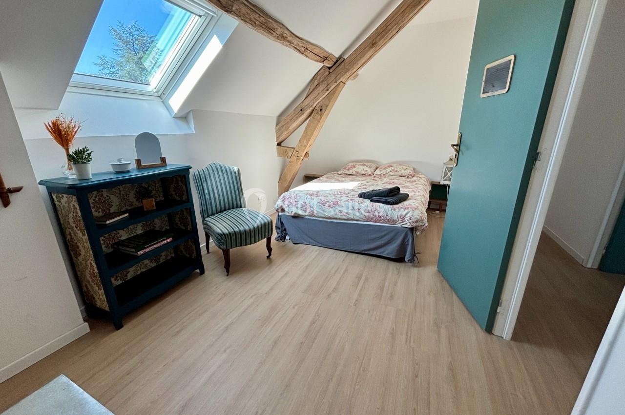 Chambre lumineuse avec lit double, poutres apparentes, fauteuil, étagère et fenêtre de toit.