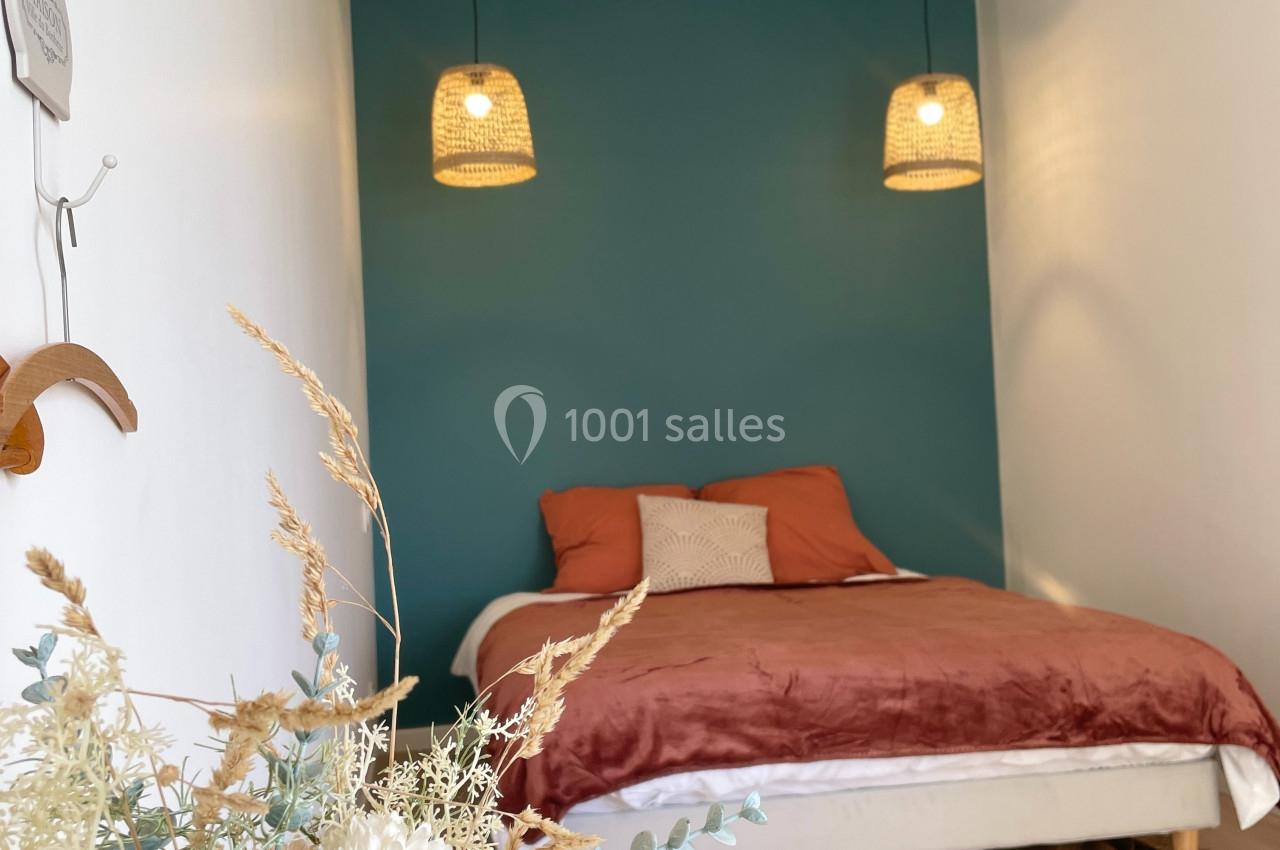 Lit double avec coussins et couverture dans une chambre minimaliste, mur bleu, suspensions en rotin et décoration florale.