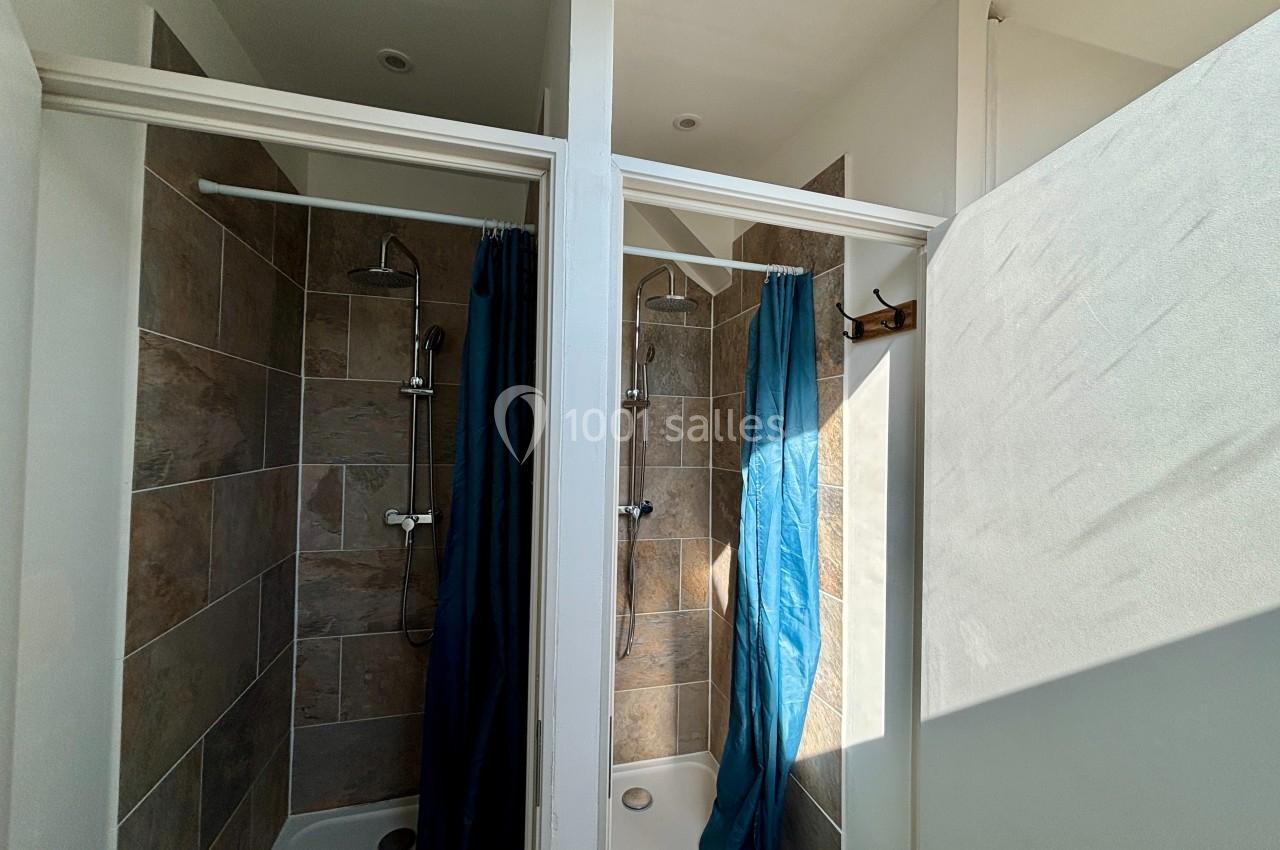 Deux douches individuelles avec rideaux bleus, carrelage beige et séparation blanche dans une pièce lumineuse.