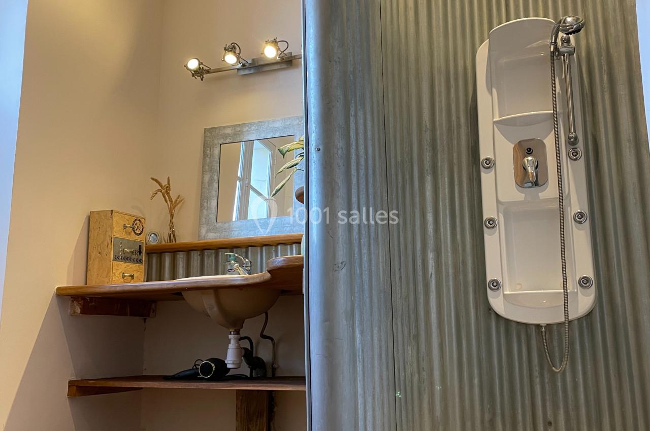 Salle de bain avec douche en tôle ondulée, lavabo sur planche en bois et miroir encadré, éclairage au-dessus.
